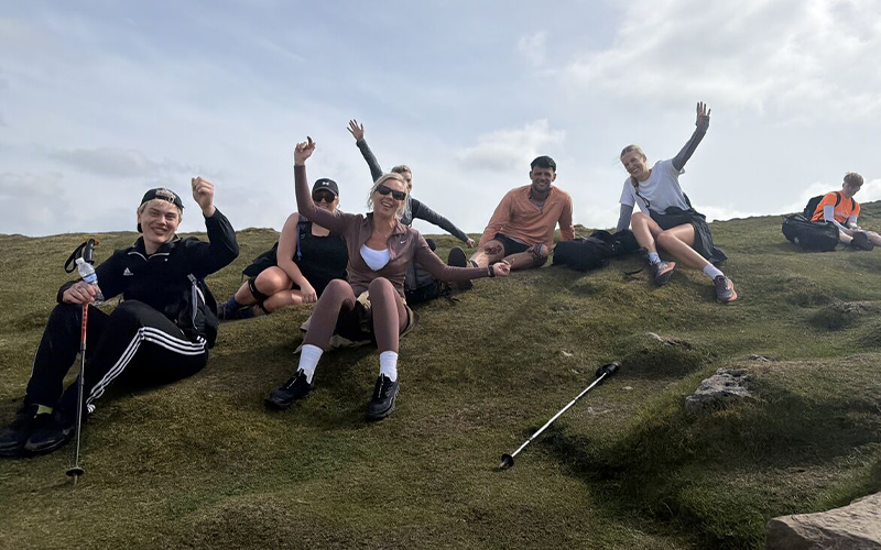 Exhausted but smiling Major Recruitment team sitting together and waving from the peak during the Yorkshire Three Peaks Challenge after a long charity hike